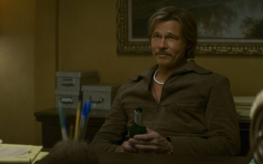 "A man with a mustache sits at a desk in a warmly lit office, holding a green bottle. The room has a lamp, filing boxes, and papers, with a painting and organized supplies in the background."