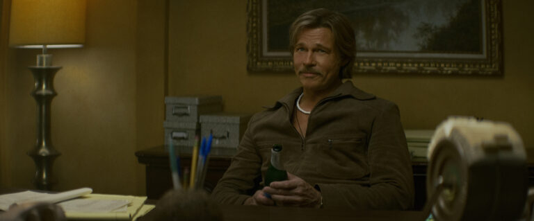 "A man with a mustache sits at a desk in a warmly lit office, holding a green bottle. The room has a lamp, filing boxes, and papers, with a painting and organized supplies in the background."